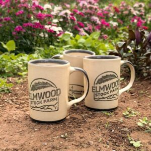 Three stoneware mugs with the Elmwood logo on them, sitting in a flower patch.