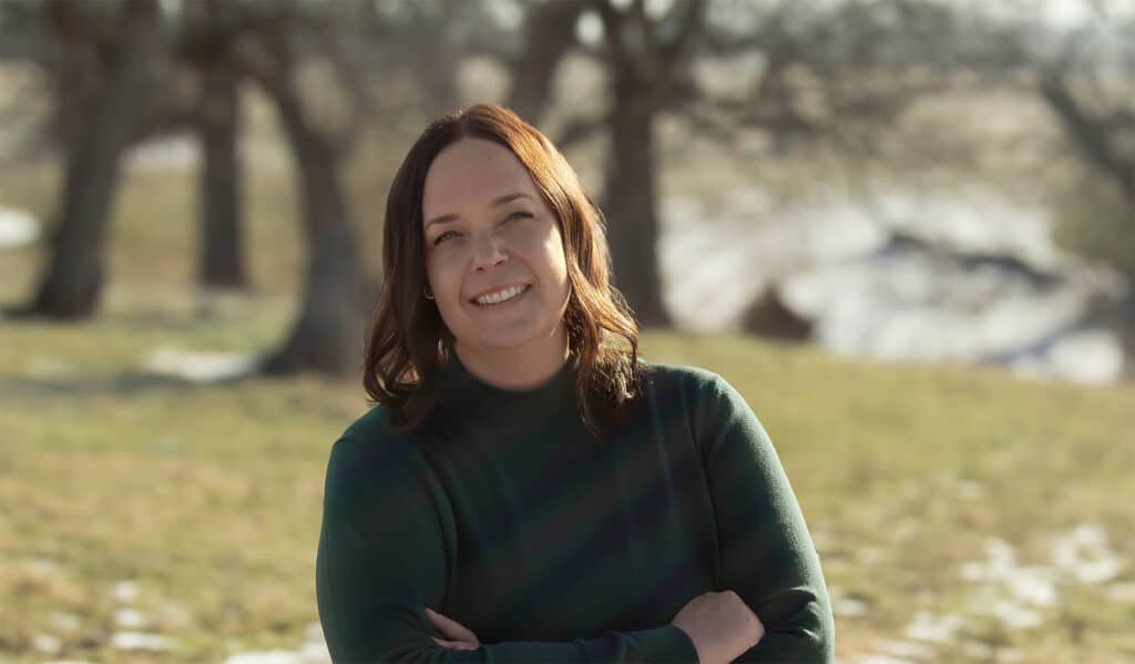 A portrait of Heidi Franklin. She has short brown hair, is wearing a green sweater, and is standing in a partially snowy field.